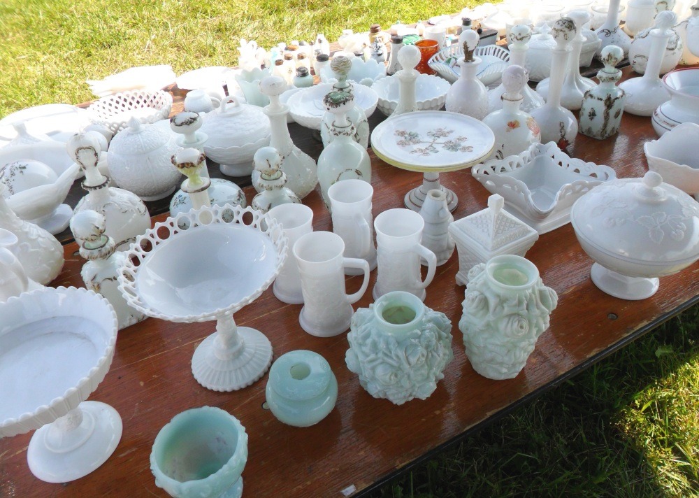 More classic antique and collectible Milk Glass pieces at an outdoor flea market in 2018, including compotes, vases, candy dishes, beer steins and stoppered bottles. 