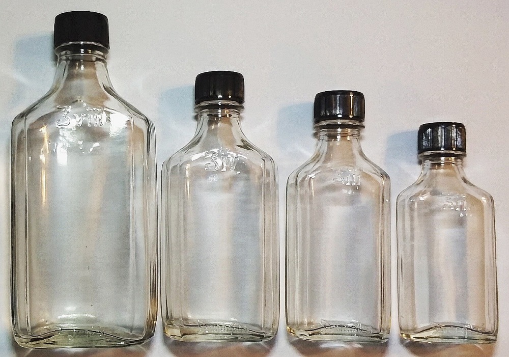 Set of four graduated size clear glass bottles with the DURAGLAS brand name embossing on them, with original black plastic lids. Made by Owens-Illinois Glass Company.