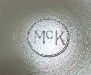 "McK in circle" mark, as it appears on the base of an ivory (custard glass) child's mug lettered "Tom & Jerry" on the front. This probably dates from the 1930s.