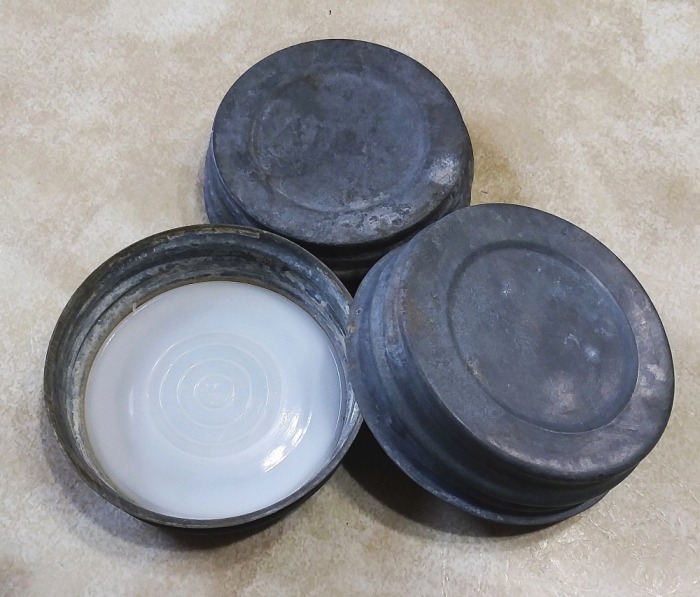 Typical zinc Mason jar lids with milk glass discs in place. The upper one is marked "ATLAS" on the front.  The one showing the disc (on lower left) is marked "BALL" on the front of the lid. The lid on right is entirely unmarked on top.