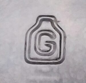 "G inside outline of a jar or bottle", logo used by Gayner Glass Company. This mark appears on a clear base shard, apparently of a wine bottle, uncertain date but perhaps 1950s-1970s era. "G inside outline of a jar or bottle". This mark appears on the base shard from what was probably a wine bottle in clear glass, perhaps 1950s-1970s era.